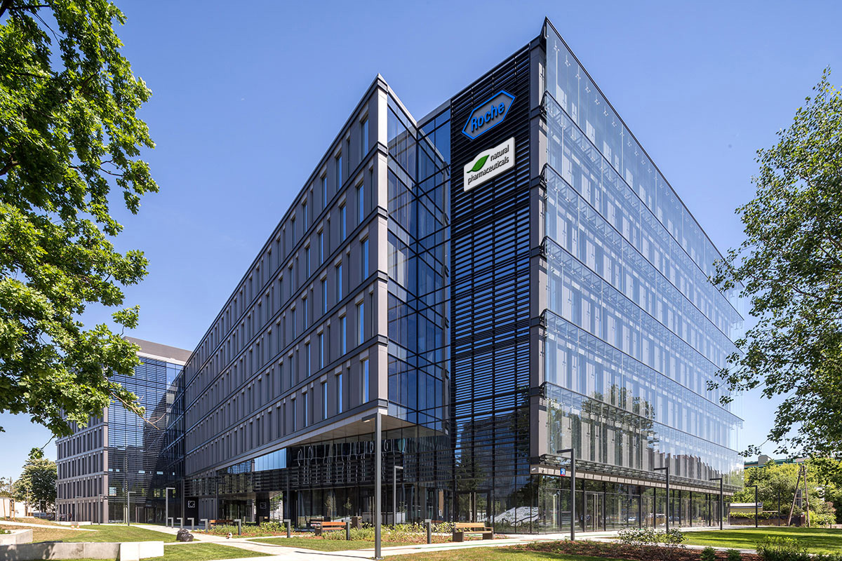 Natural Pharmaceuticals VitaSolaris® D3 Moderne klaasfassaadiga Roche ja Natural Pharmaceuticals kontorihoone päikeselises keskkonnas, roheliste puude lähedal.