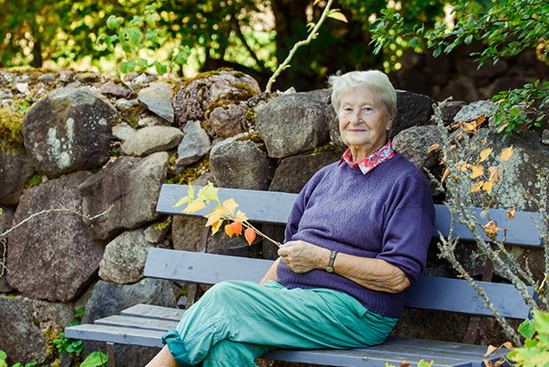 Natural Pharmaceuticals VitaSolaris® D3 Eakas naine istub pingil kivimüüri ees, hoides sügislehtedega okst looduses rahulikult.